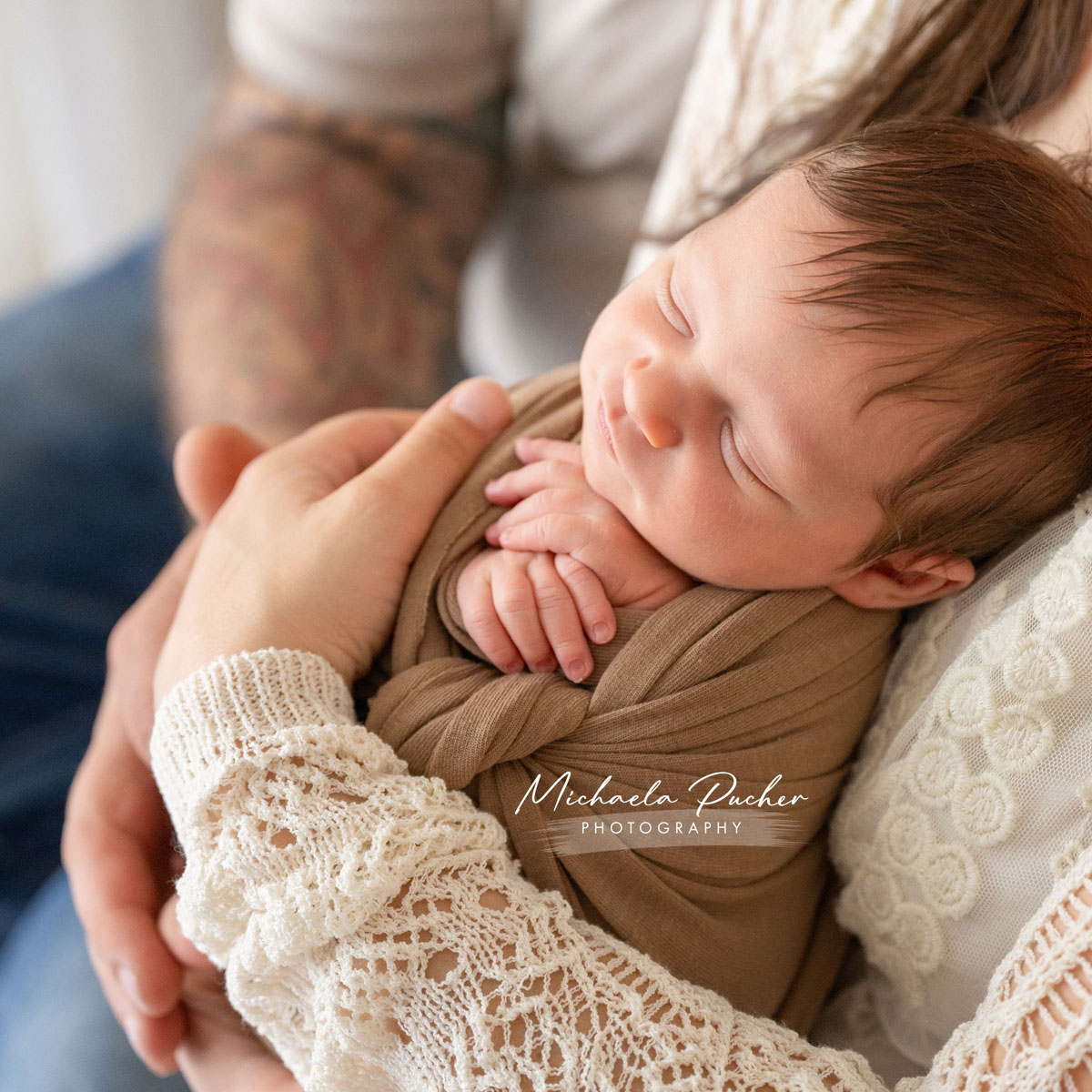 Schlafendes Neugeborenes liegt eingewickelt in braune Tücher in den Armen seiner Eltern; die Hände der Eltern halten das Baby behutsam.